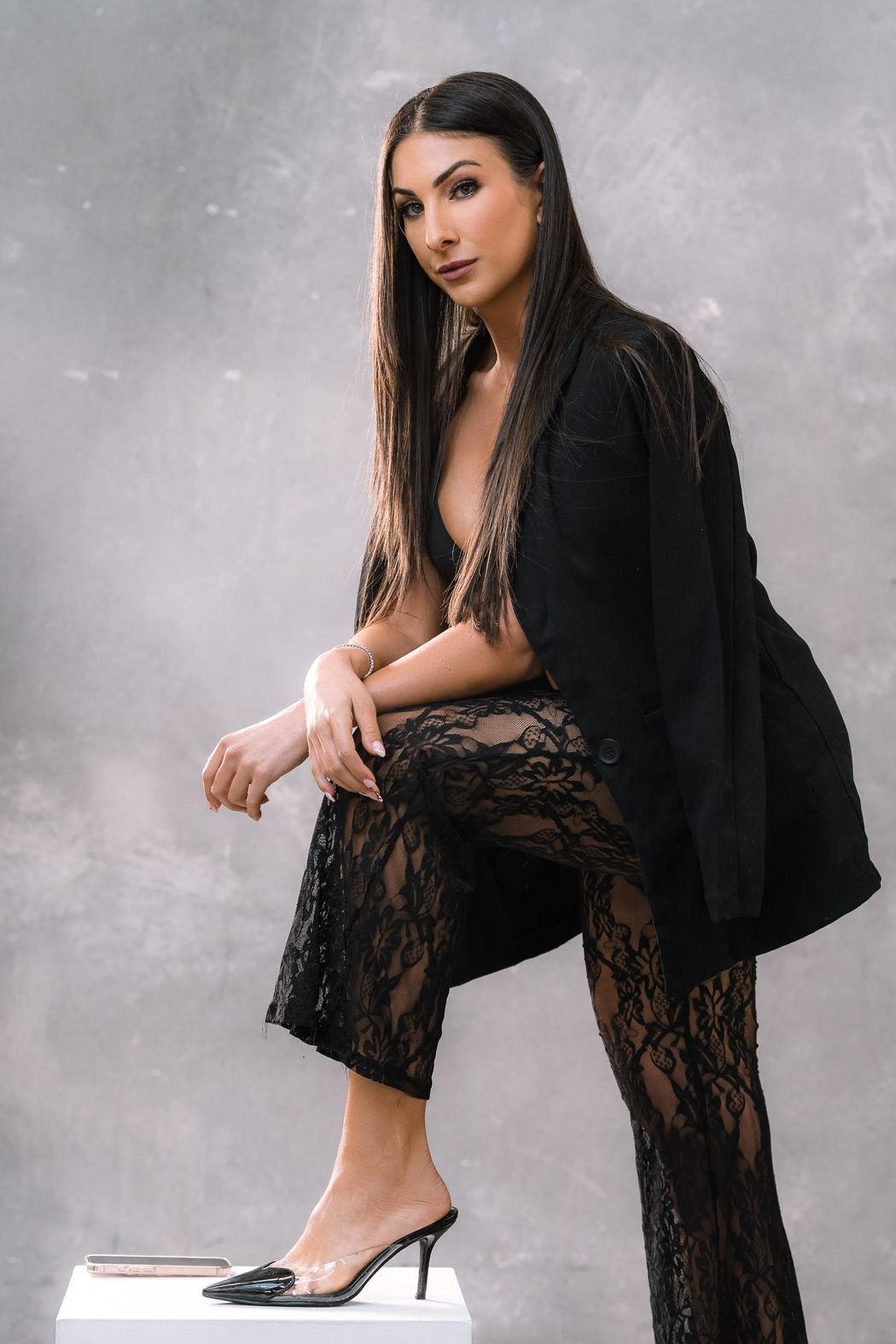 Woman in a black blazer and lace trousers, foot raised on a white block
